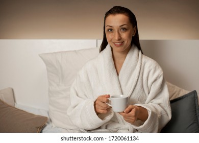 Attractive Woman In Bathrobe Sitting In Bed Looking With Smile At Camera While Drinking Hot Herbal Tea. Young Brunette Woman Resting At Home In Leisure Time. Happy Girl Relaxing Before Bedtime