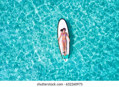 Attractive woman in bikini is sunbathing on a surfboard, aerial view