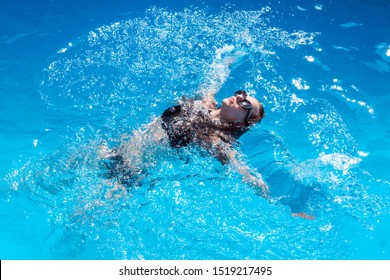 Attractive Woman In A Black Bathing Suit Floating On Her Back In The Swimming Pool And Relaxing, With Copy Space. View From The Top. Young Female Athlete Swims On Her Back In The Blue Water Of Pool