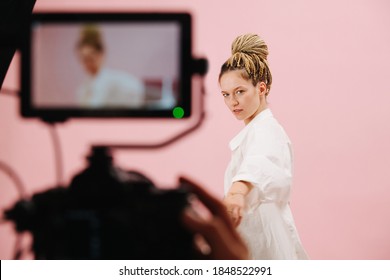 Attractive woman with blond afro braids in a bun posing in front of a camera with a field monito. Over pink background. Blurred equipment in a foreground.