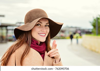 Attractive woman in a brown hat and beige coat looking at the camera points the finger at you and smiles. Pick me