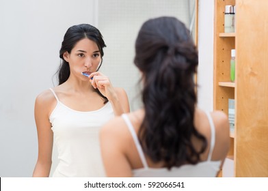 Attractive Woman Brushing Teeth In Bathroom. Young Woman Brushing Teeth While Looking At Mirror. Girl Brushing Teeth Before Going To Bed At Night.