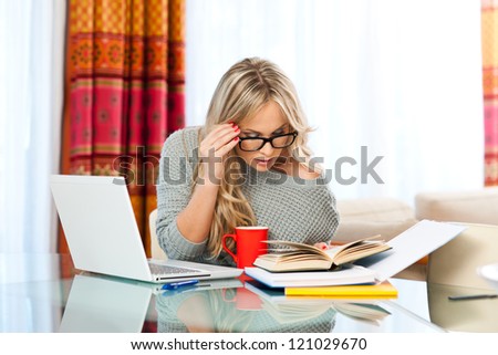 attractive woman with cup of coffee, book and laptop sitting at her home desk and working
