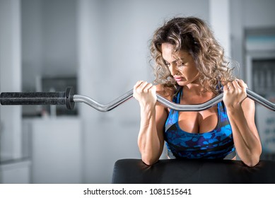 Attractive Woman With Curly Hair Doing Preacher Curl Biceps Exercise On Bench With EZ Curl Bar In Modern Fitness Center. Toned Image.