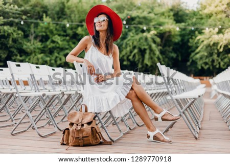 attractive woman dressed in white dress, red hat, sunglasses sitting in summer open air theatre on chair alone, spring street style fashion trend, accessories, traveling with backpack