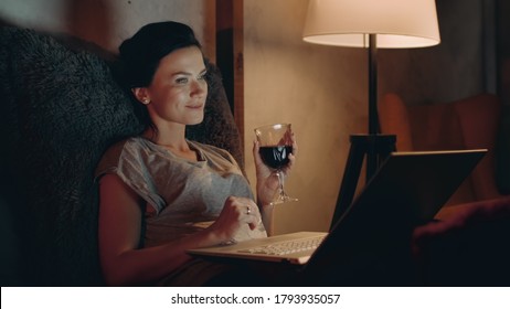 Attractive Woman Drinking Red Wine With Notebook In Night Room. Closeup Smiling Woman Relaxing With Glass Of Wine At Home. Pretty Girl Looking Laptop With Wineglass On Couch.