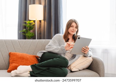 Attractive woman, freelancer holding and using digital tablet, sitting at cozy sofa in living room. Successful businesswoman text message in home. Online technology concept, remote job - Powered by Shutterstock