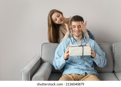 Attractive Woman Gives A Gift To A Man. A Young Couple In Love And Congratulations On The Holiday. Isolated On Bright Background At Home. Close And Open Eyes. Surprise.