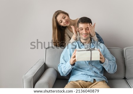 Attractive woman gives a gift to a man. A young couple in love and congratulations on the holiday. Isolated on bright background at home. Close and open eyes. Surprise.