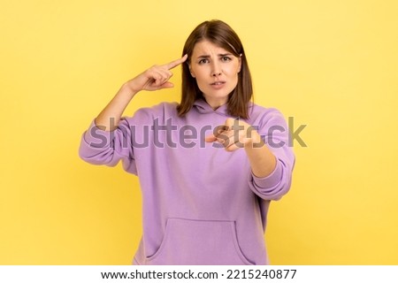  Attractive woman with hair holding finger near temple and pointing on you, showing dumb gesture, wearing purple hoodie. Indoor studio shot isolated on yellow background.