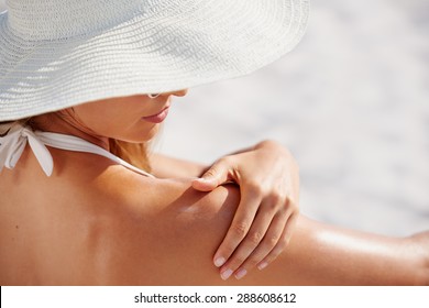 Attractive woman with healthy skin applying sunscreen to shoulder wearing white sun hat