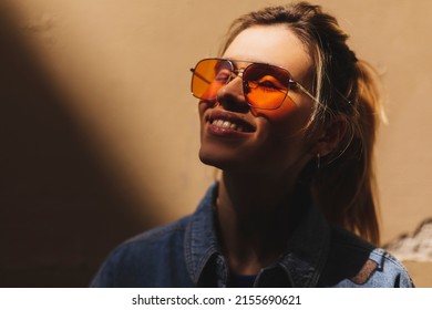 Attractive Woman With High Ponytail Standing Near Beige Wall In Old City. Girl Joy Of Sun Light, Wear Orange Sunglasses, Closed Eyes. Shadow And Light Of Sunny Summer, Spring Or Fall Day.