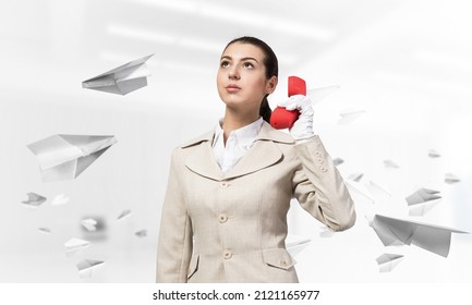 Attractive Woman Holding Vintage Red Phone In Office With Flying Paper Planes. Elegant Operator In White Business Suit Posing With Landline Phone. Hotline Telemarketing And Business Communication.