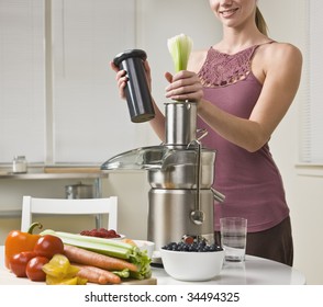 Attractive Woman With Juicer Machine, Adding Celery And Smiling. Horizontal.