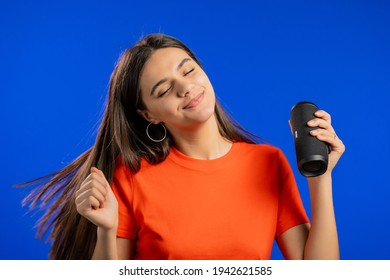 Attractive Woman Listening To Music By Wireless Portable Speaker - Modern Sound System. Lady Dancing, Enjoying On Blue Studio Background. She Moves To The Rhythm Of Music.