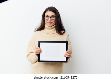  Attractive Woman With Long Dark Hair, Smiling Cutely Looking At The Camera, Standing In A Sweater With A Black Frame In Her Hands On A Light Background. Studio Photography With Space For Mockup