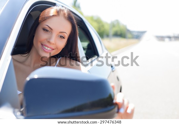 Attractive woman\
is looking at side mirror of her car and adjusting it with her\
hand. The lady is smiling with satisfaction. She is sitting at seat\
of driver. Copy space in right\
side