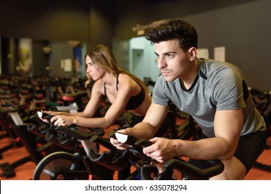 Attractive Woman And Man Biking In The Gym, Exercising Legs Doing Cardio Workout Cycling Bikes. Couple In A Spinning Class Wearing Sportswear.