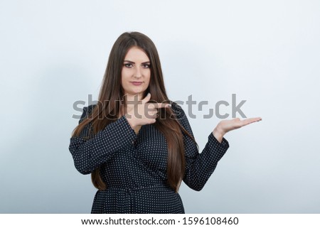 Attractive woman over isolated gray background wearing casual black dress with white polka dot, pointing fingers aside, looking at camera. People lifestyle concepte.