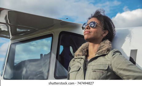 Attractive woman pilot wearing sunglasses standing in the sunshine on an airfield resting against her small private airplane