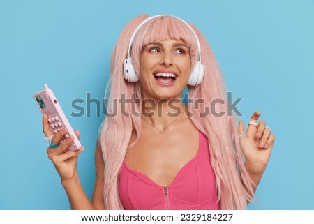 Attractive woman with pink hair and headphones wearing Barbie style pink top posing on blue background with phone in hand and smiles, vintage fashion concept, copy space