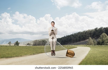 Attractive woman playing trumpet brass on asphalt road. Young businesslady in white business suit and gloves sitting on big light bulb with music instrument. Musician practicing and performing outdoor - Shutterstock ID 2256850965