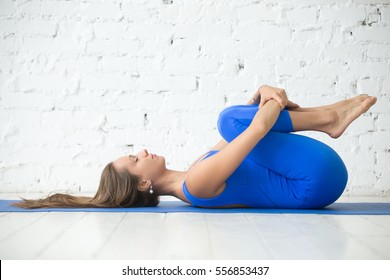 Attractive Woman Practicing Yoga, Knees To Chest Exercise, Apanasana Pose, Working Out, Wearing Sportswear, Blue Suit, Indoor Full Length, White Loft Studio Background. Tattoo On Foot 