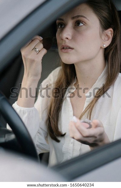 An attractive woman\
puts a blush with a large brush while sitting behind the wheel of a\
car. Early morning, a businesswoman is late for work. Toning powder\
for the face