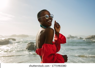 Привлекательная женщина в красном платье танцует на пляже. Африканская женщина в красном сарафане и солнцезащитных очках развлекается на пляже.