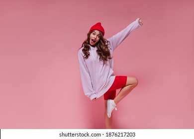 Attractive Woman In Red Hat, Cycling Shorts And Purple Hoodie Moves On Pink Background. Curly Girl Rises Leg On Isolated Backdrop.