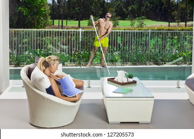 Attractive Woman Relaxing In Backyard Patio With Pool Boy Cleaning The Pool