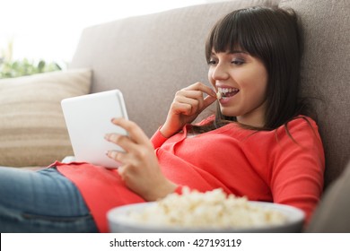 Attractive woman relaxing on the couch at home, she is watching videos online and eating popcorn, movies streaming concept
