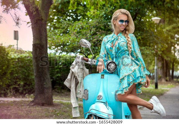 Attractive woman riding on\
motorbike in street, summer vacation style, traveling, smiling,\
happy, having fun, stylish outfit, adventures. Girl on a blue\
scooter - Image