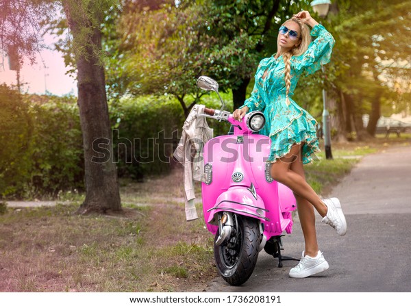 Attractive woman riding on\
motorbike in street, summer vacation style, traveling, smiling,\
happy, having fun, stylish outfit, adventures. Girl on a pink\
scooter - Image