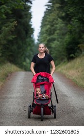 Attractive Woman Running On A Trail Pushing Her Baby Stroller Outdoors. Vertical Image Of A Fit Female Working Out While Being An Active Mom