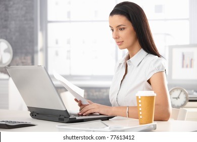 Attractive woman sitting at desk in office, working with laptop computer, holding document, having takeaway coffee.? - Powered by Shutterstock