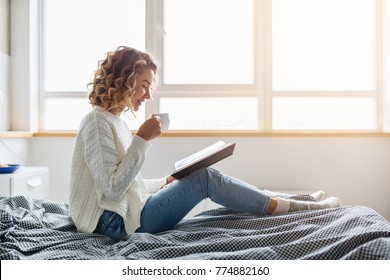 attractive woman sitting on bed in the morning, drinking tea, reading book, curly hair, casual style, blue jeans, white sweater, feeling comfortable at home, having rest, smiling - Powered by Shutterstock