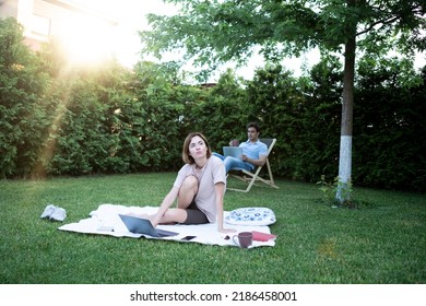 Attractive Woman Sitting On Picnic Blanket On The Lawn In The Backyard With Her Husband Working On Laptop On Background