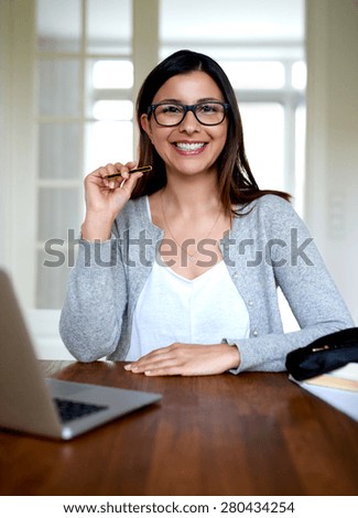 Attractive woman sitting upright at desk in home office smiling and holding pencil.