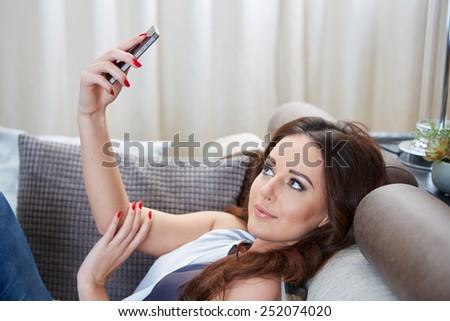 Attractive woman smiling as she reads an sms message on her mobile while relaxing barefoot on a sofa in her living room