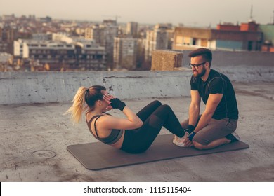 Attractive woman in sportswear doing sit ups on a building rooftop terrace with the assistance of a personal fitness instructor. Focus on the instructor - Powered by Shutterstock