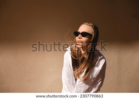 Attractive woman standing near beige wall in old city. Girl joy of sun light, wear black sunglasses and white shirt look at sun. Shadow and light of sunny summer. Wind blowing hair, woman laughing.