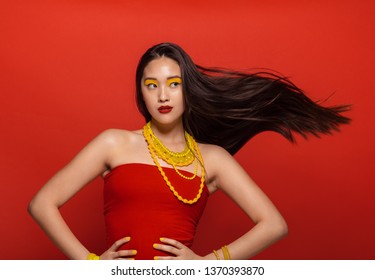 Attractive woman with stylish stage make up and hair flying on red background. Asian female model with creative make up and accessories.