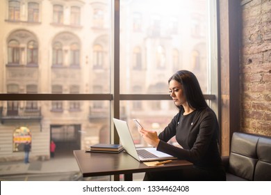Attractive woman successful commercial real estate analyst checks the status of the account on smartphone while sitting with laptop computer in coffee shop near window with copy space. Online banking 