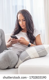 Attractive woman in sweatpants and t-shirt texting on a phone in a white light background of her apartment.