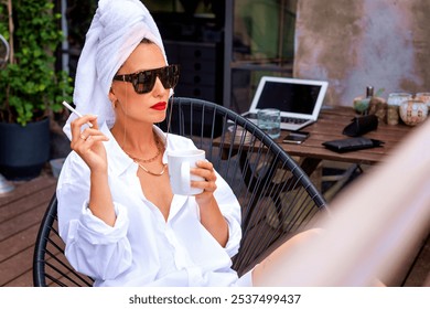 Attractive woman with towel turban on head wearing sunglasses while having coffee on balcony and smoking cigarette in the morning. - Powered by Shutterstock