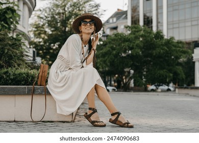 attractive woman walking in street on vacation dressed in white summer fashion dress straw hat sunglasses and purse, smiling happy having fun, stylish accessories trend