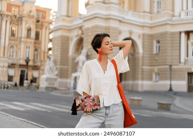 attractive woman walking in street in summer fashion trend style outfit smiling happy, white blouse, orange bag, purse, earrings, city center Europe vacation, positive mood