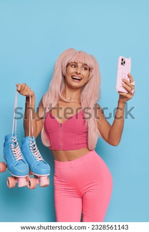 Attractive woman wearing Barbie style top and leggins posing on blue background with roller-skates and phone in hands and takes selfie, vintage fashion concept, copy space