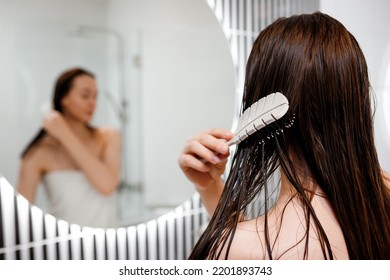 Attractive woman in white towel with comb brushing her wet hair after showering at home in front of bathroom mirror. Cares about healthy and clean hair. Beauty concept - Shutterstock ID 2201893743